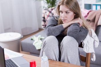 Sad and tired woman with postpartum pain working beside table, looking on laptop, sitting in messy room